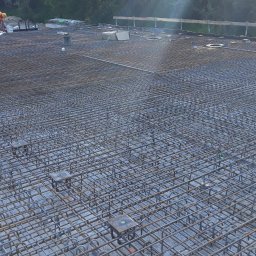 DREBEKO - Zbrojenie płyty fundamentowej z widocznym pracownikiem w pomarańczowym kombinezonie. Siatka z prętów stalowych, podkładki dystansowe. Etap budowy domu.