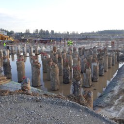 DREBEKO - Betonowe pale fundamentowe wystające z wody na placu budowy, widoczny dźwig i pracownicy w odblaskowych ubraniach. W tle drzewa i niebo.