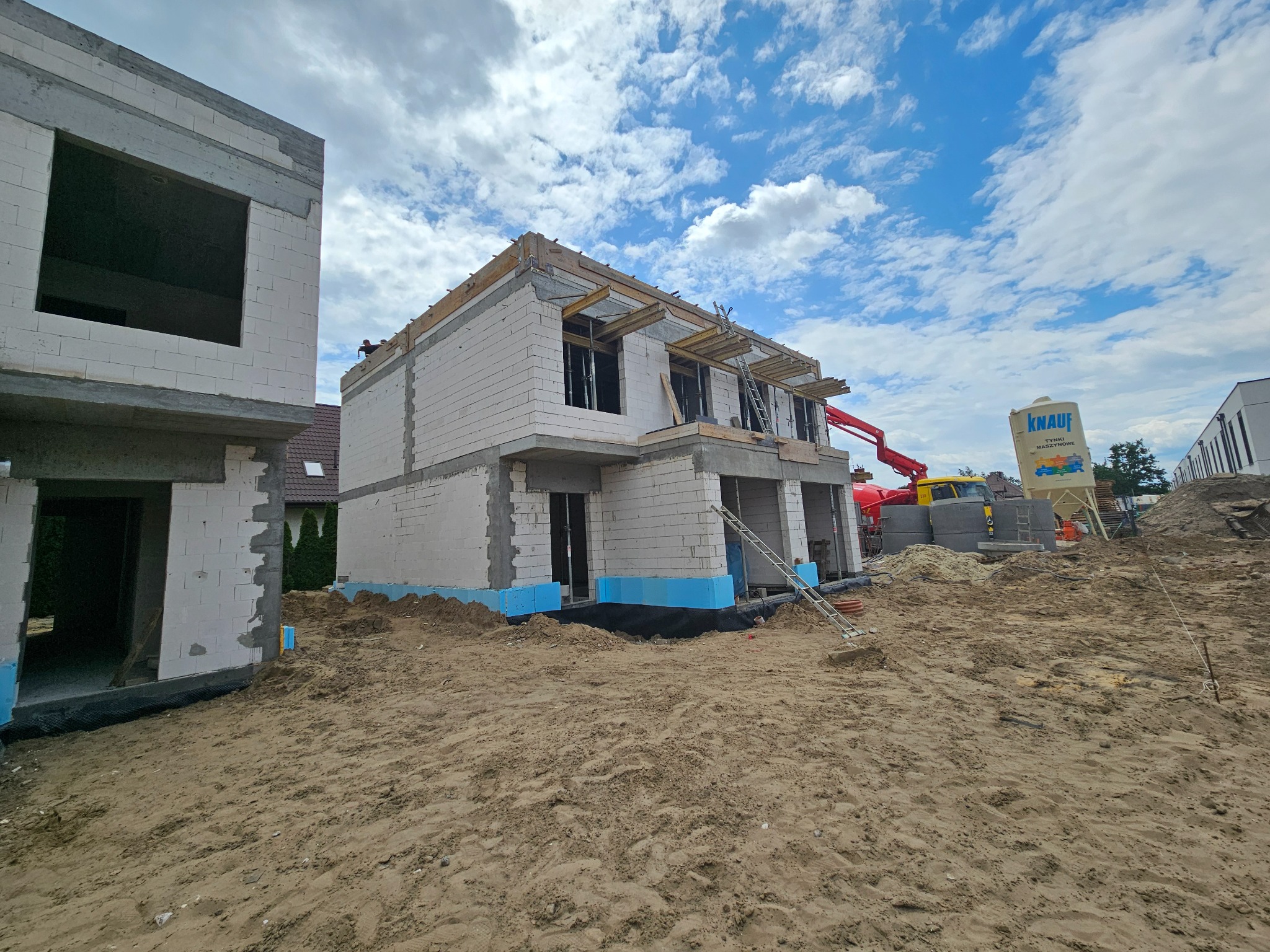 Budowa domu z białych bloczków, widoczny dźwig i materiały budowlane. Niebieska izolacja fundamentów. Częściowo zrealizowany stan surowy otwarty.