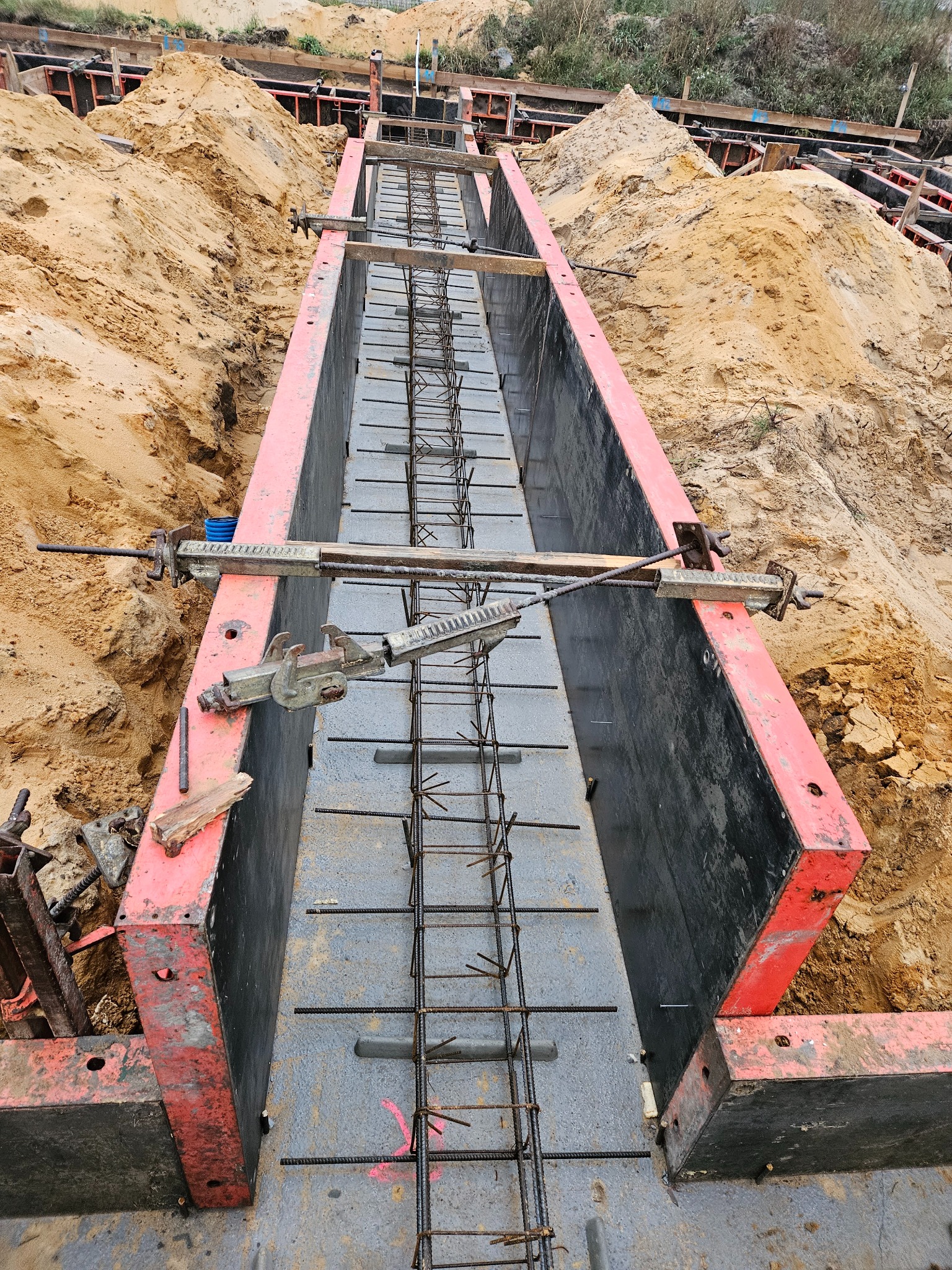 Zbrojenie ławy fundamentowej w szalunku, widok z góry. Stalowe pręty tworzą siatkę wewnątrz formy, przygotowane do zalania betonem. Czerwono-czarne szalunki otoczone ziemią.