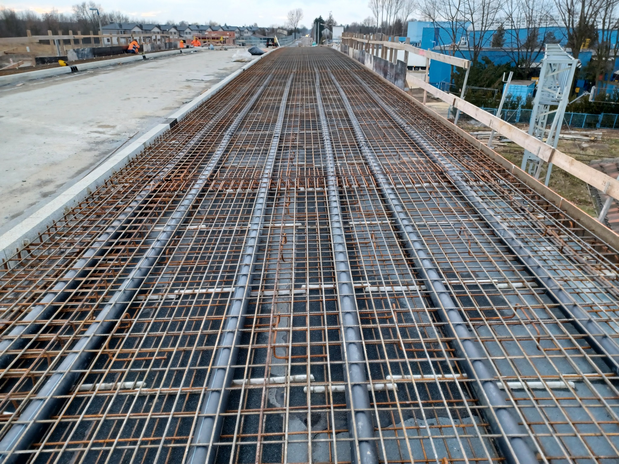 Zbrojenie płyty fundamentowej z rurami instalacyjnymi, widok z góry. Na drugim planie budynki w trakcie budowy i pracownicy w pomarańczowych ubraniach.