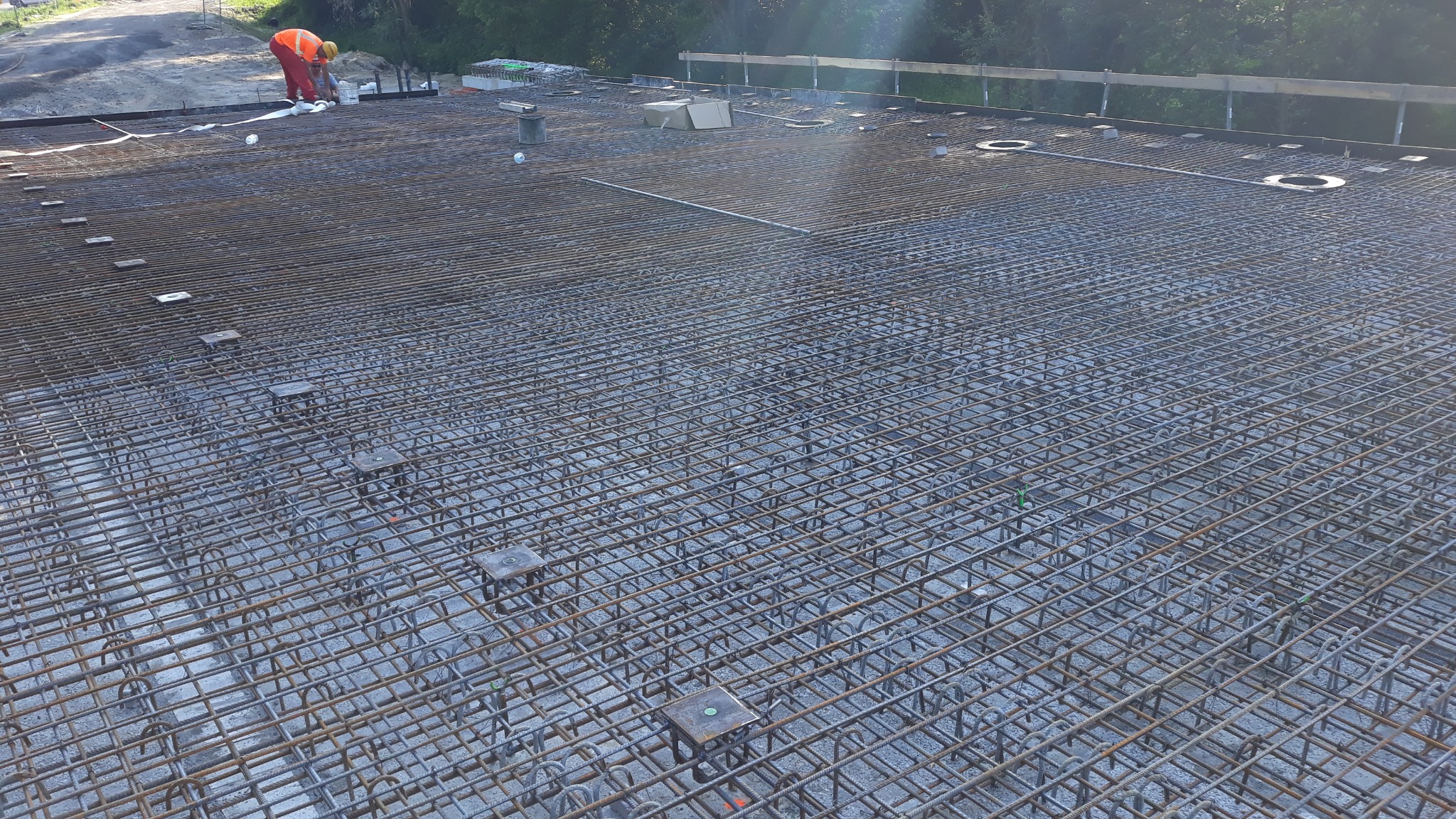 Zbrojenie płyty fundamentowej z widocznym pracownikiem w pomarańczowym kombinezonie. Siatka z prętów stalowych, podkładki dystansowe. Etap budowy domu.