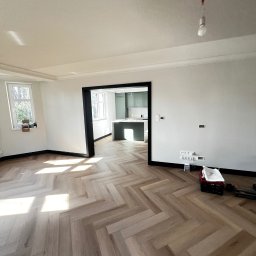 Katruk Technologies - Jasne parkiet w jodełkę francuską w nowo wykończonym pokoju z widokiem na kuchnię. Świeże, białe ściany z czarnymi listwami i ościeżnicami. W rogu narzędzia.