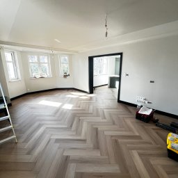 Katruk Technologies - Jasny parkiet ułożony we wzór jodełki francuskiej w przestronnym, odnowionym pokoju z białymi ścianami i czarnymi listwami przypodłogowymi. Widoczna drabina i skrzynka narzędziowa.