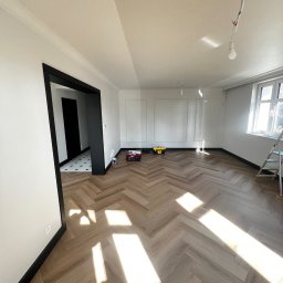 Katruk Technologies - Jasne wnętrze po remoncie: parkiet w jodełkę, białe ściany z dekoracyjnymi panelami, czarne obramowania drzwi i okien, narzędzia pracy w rogu.