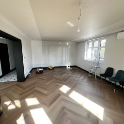 Katruk Technologies - Jasne wnętrze po remoncie: parkiet w jodełkę, białe ściany z dekoracyjnymi panelami, czarne ramy drzwi. Widoczne narzędzia i drabina.