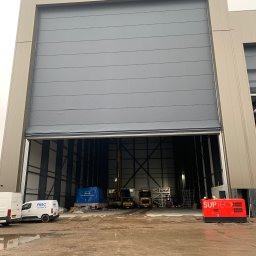 Cendre Installatie - Częściowo otwarta, przemysłowa brama segmentowa w szarym kolorze, z widocznym wnętrzem hali i pojazdami. Brama zamontowana w nowoczesnym budynku o jasnej elewacji.