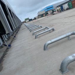 Cendre Installatie - Rząd stalowych odbojników rurowych przy rampie załadunkowej magazynu, z widocznymi dokami i pracownikami w tle. Pochmurne niebo.