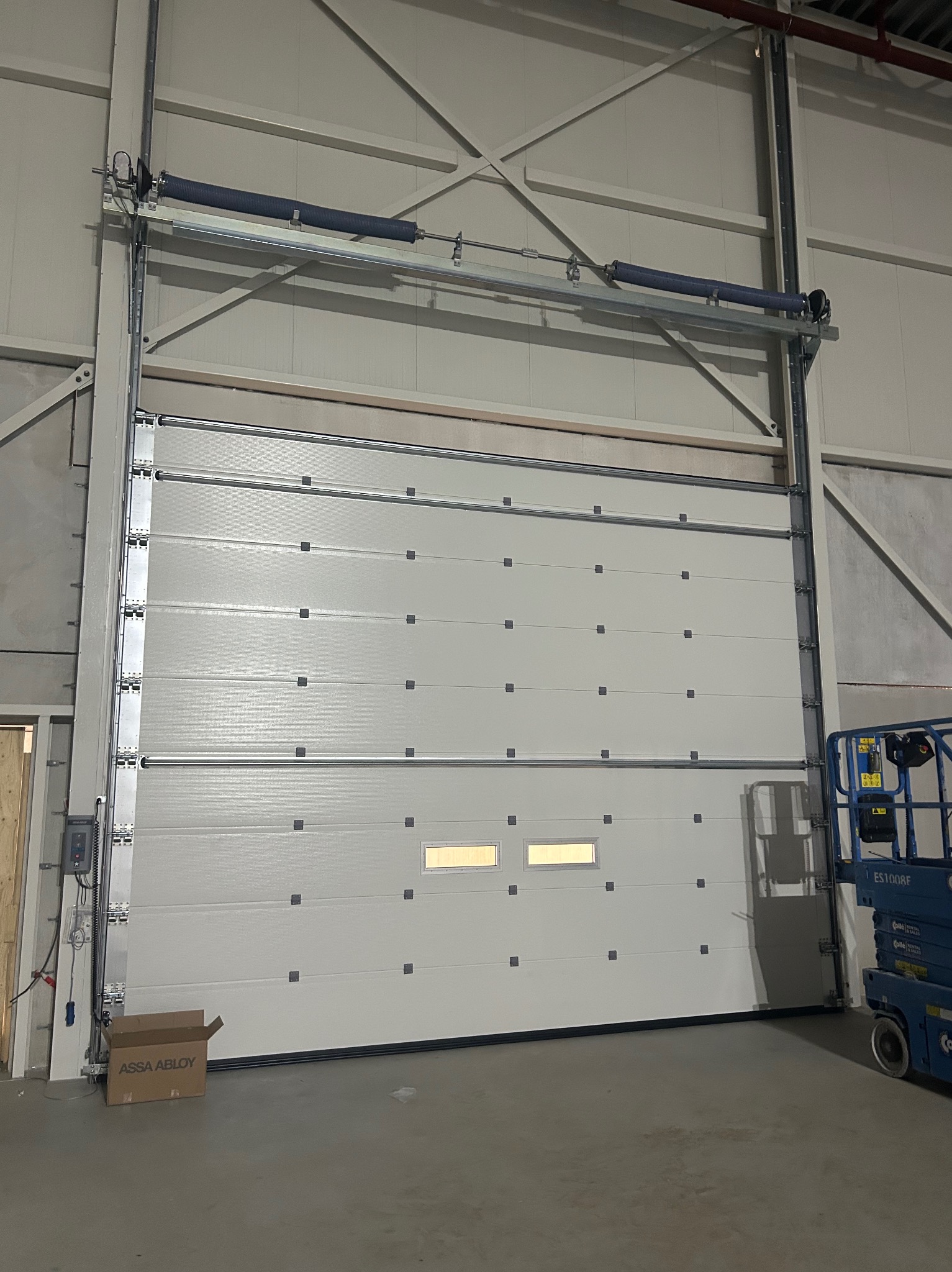 Zamknięta, segmentowa brama garażowa z dwoma okienkami, widoczna z bliska w hali w Prabutach. Obok podnośnik koszowy i karton po elementach bramy.