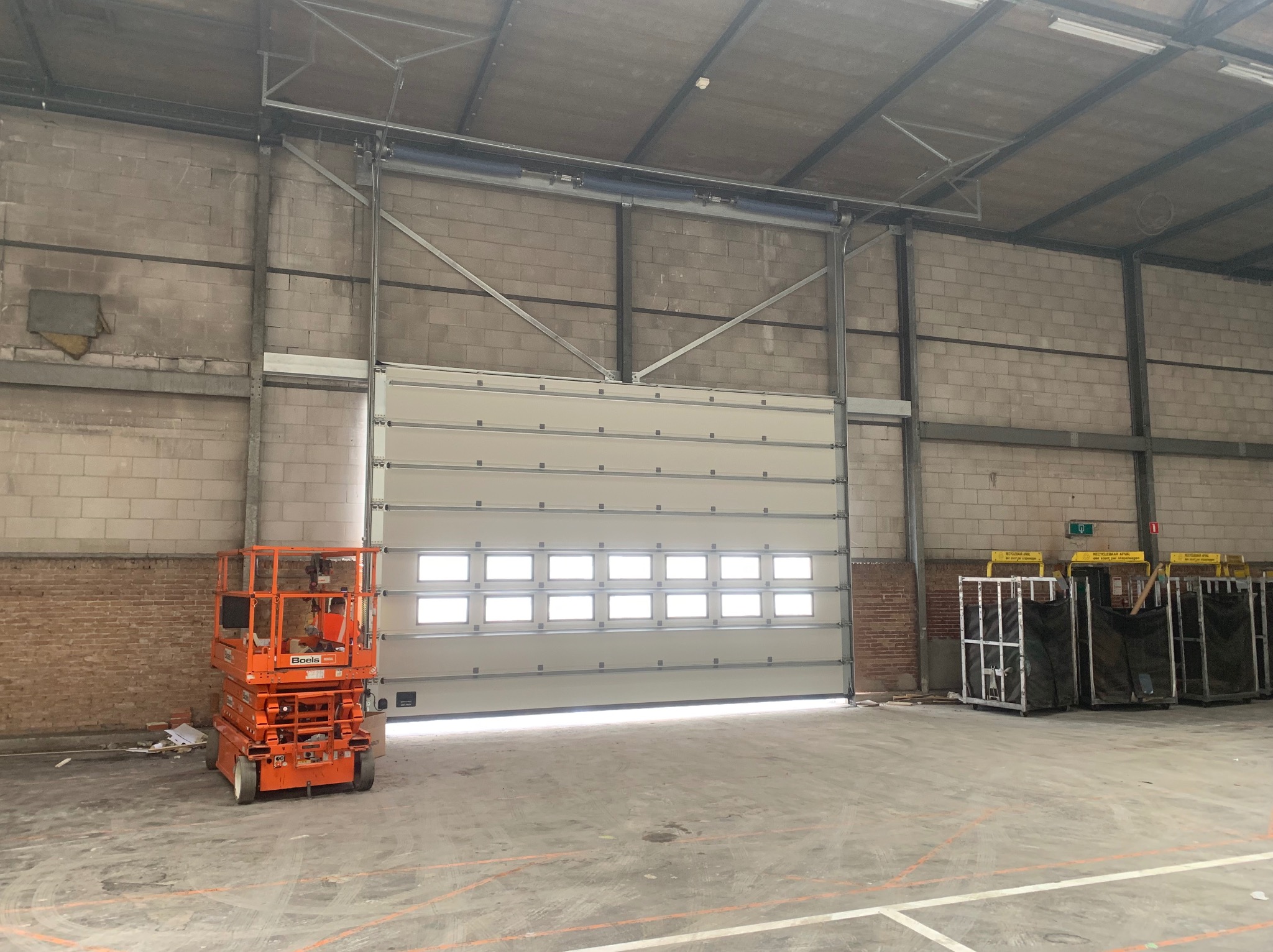 Biała segmentowa brama przemysłowa z oknami w hali, obok podnośnik koszowy Boels. Widoczne ceglane i betonowe ściany oraz stalowa konstrukcja dachu.