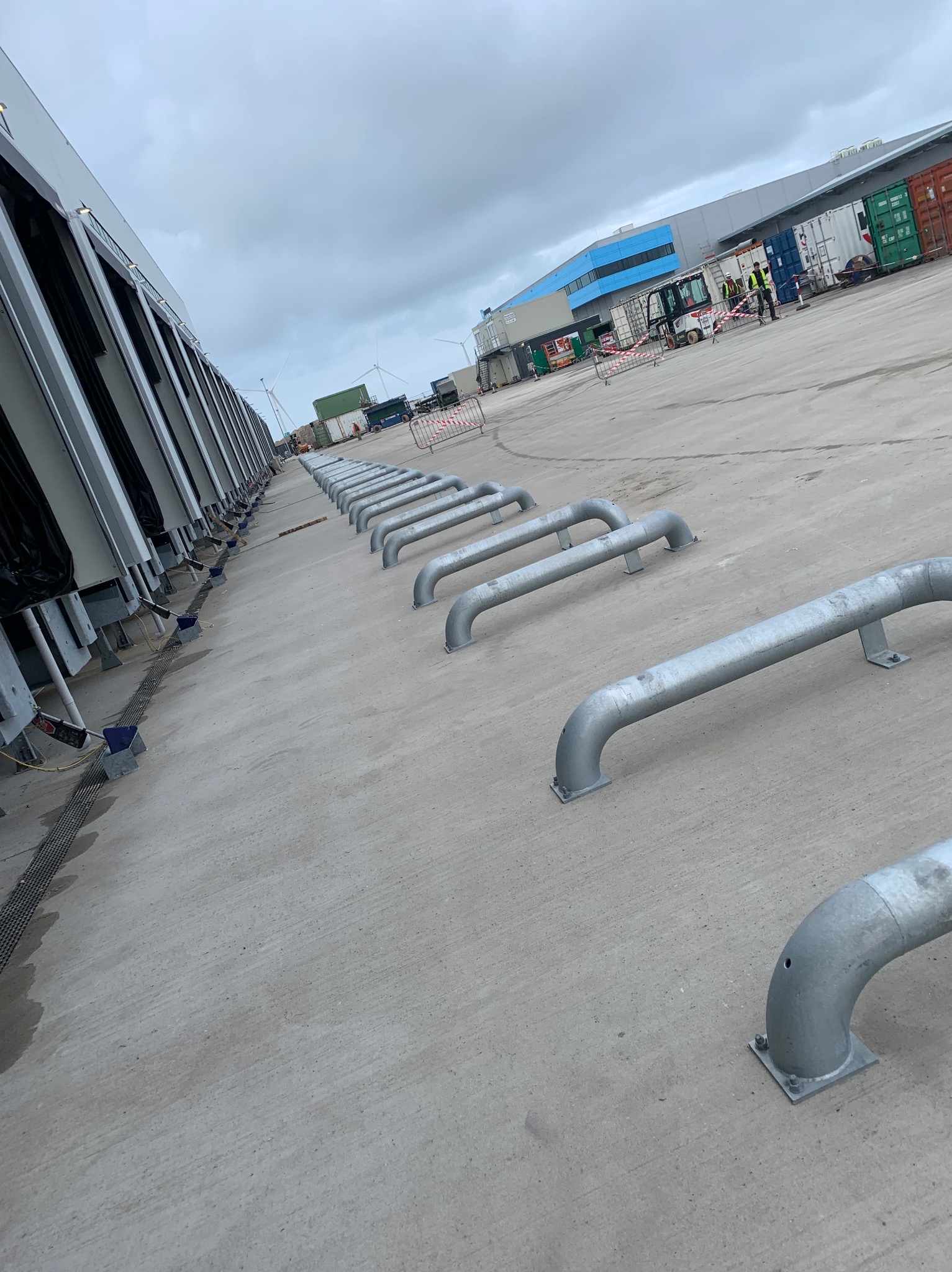 Rząd stalowych odbojników rurowych przy rampie załadunkowej magazynu, z widocznymi dokami i pracownikami w tle. Pochmurne niebo.