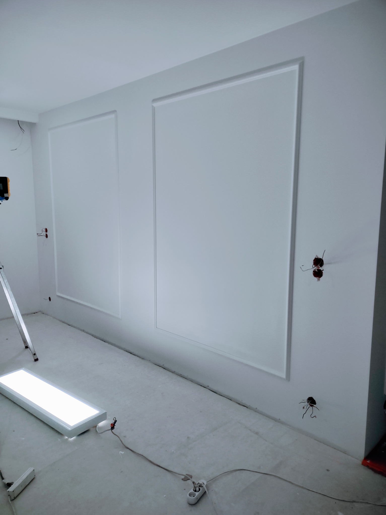 Białe ściany z dekoracyjnymi panelami w trakcie wykańczania. Widoczne kable elektryczne, drabina i lampa robocza. Minimalistyczny design wnętrza w surowym stanie.