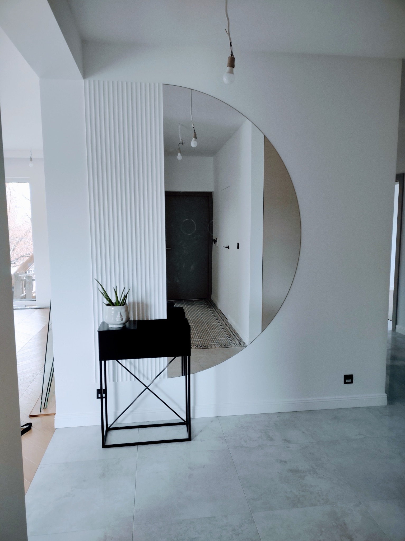 Nowoczesny hol z okrągłym lustrem, dekoracyjną ścianą z pionowymi listwami i czarną konsolą z rośliną. Minimalistyczny design, jasne kolory, płytki podłogowe.