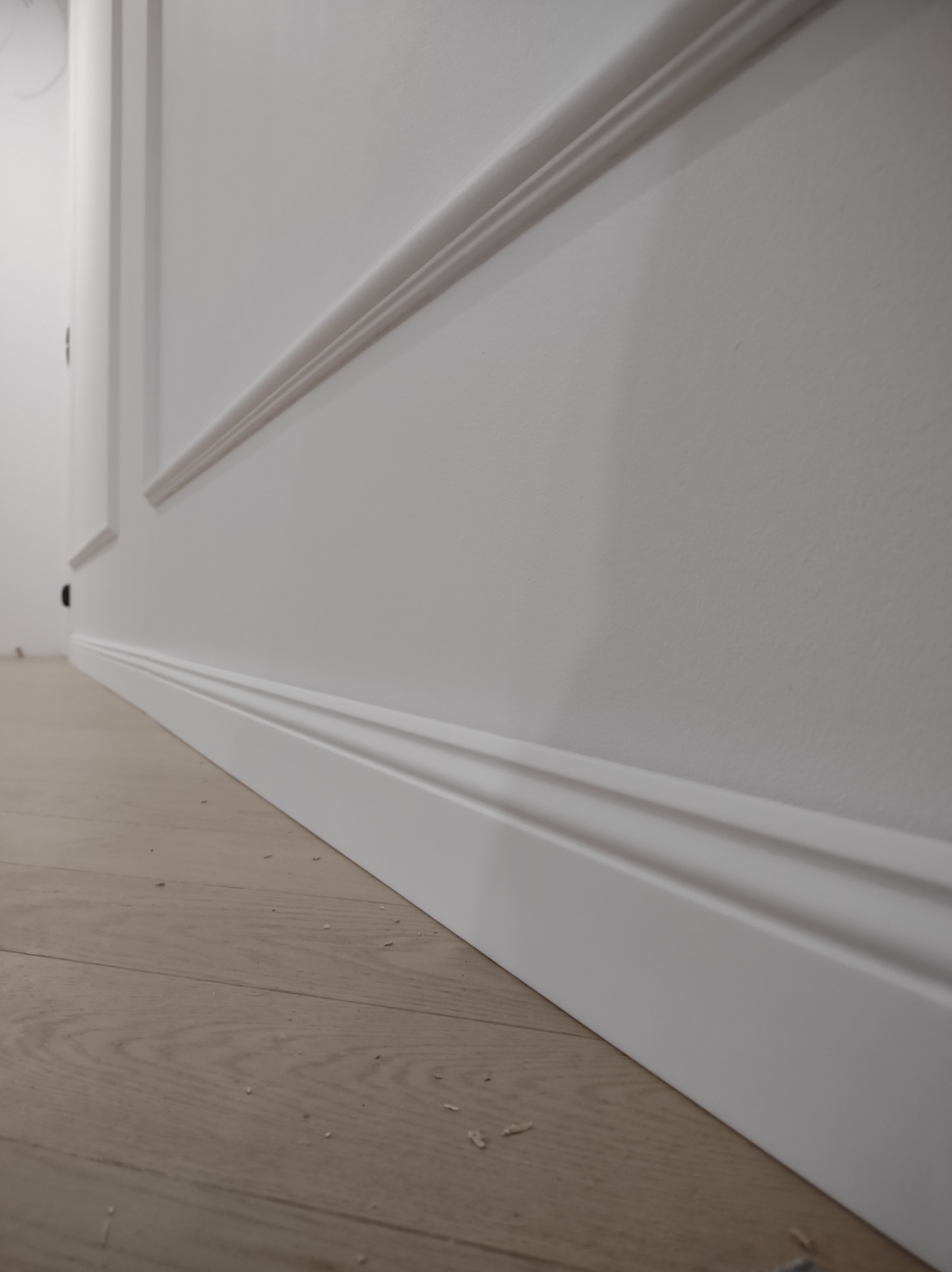 Biała ściana z dekoracyjnymi listwami przypodłogowymi i sztukaterią, widok z dołu, podkreślający detale wykończenia wnętrza i połączenie ściany z podłogą.