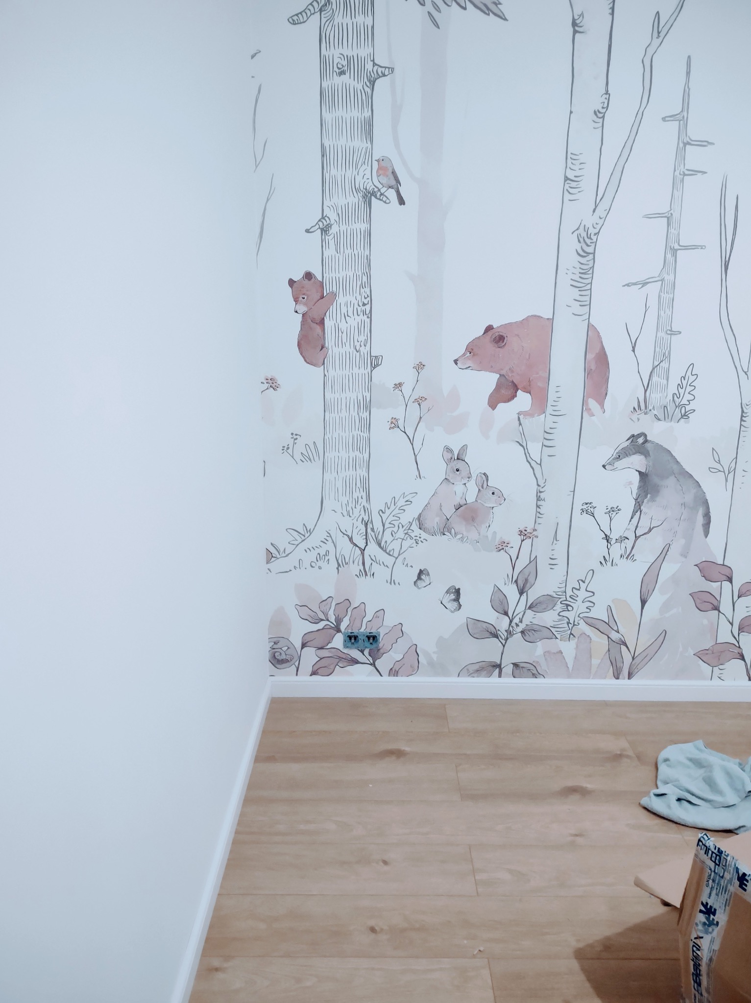 Pokój po tapetowaniu z motywem leśnych zwierząt: niedźwiedzie, zające, borsuk. Jasne panele podłogowe, białe ściany. Widoczne gniazdko elektryczne.
