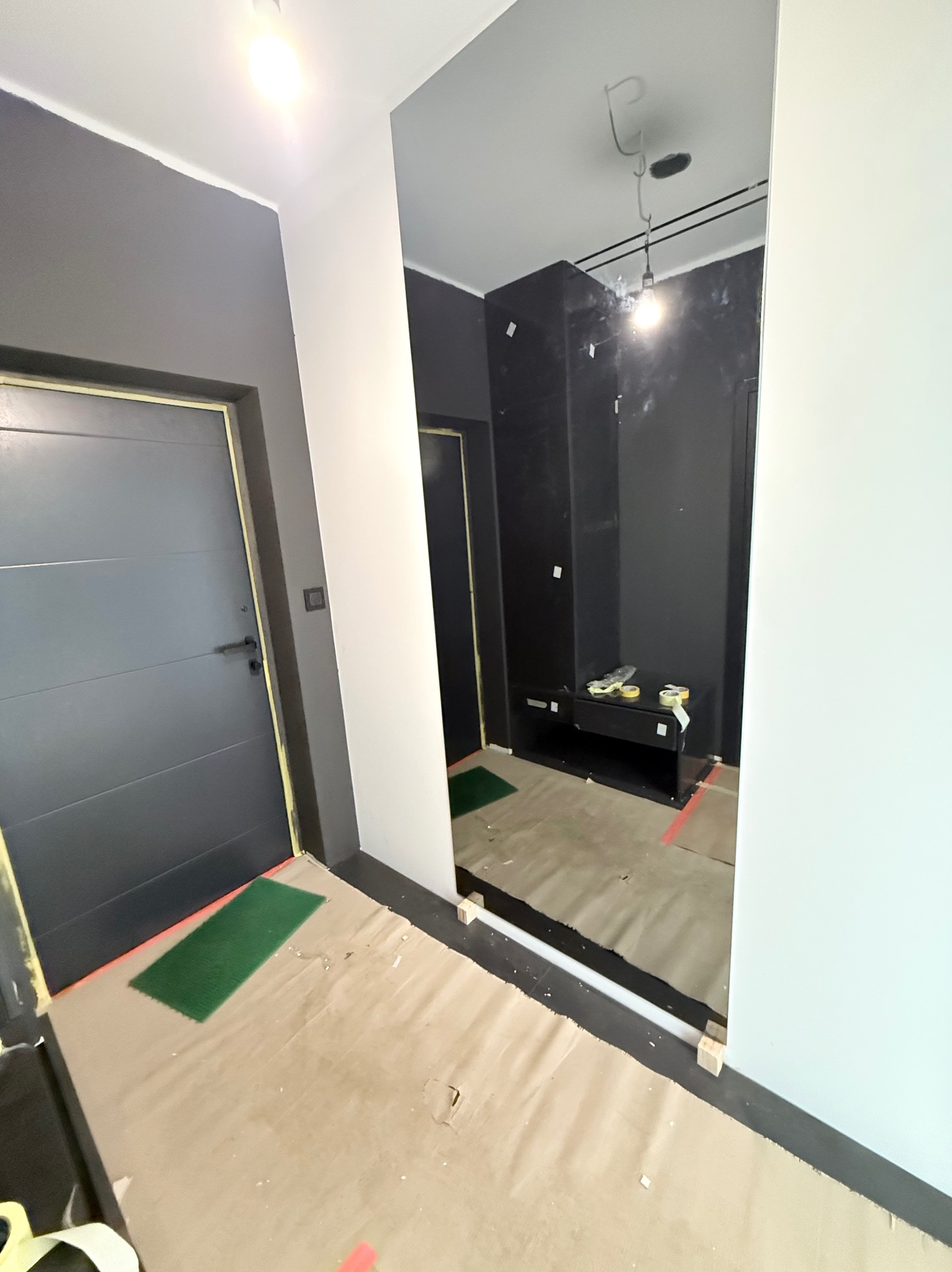 Nowoczesny przedpokój w trakcie prac: szara ściana z drzwiami, duże lustro na ścianie, zabezpieczona podłoga. Widoczne przygotowania do dalszych prac.