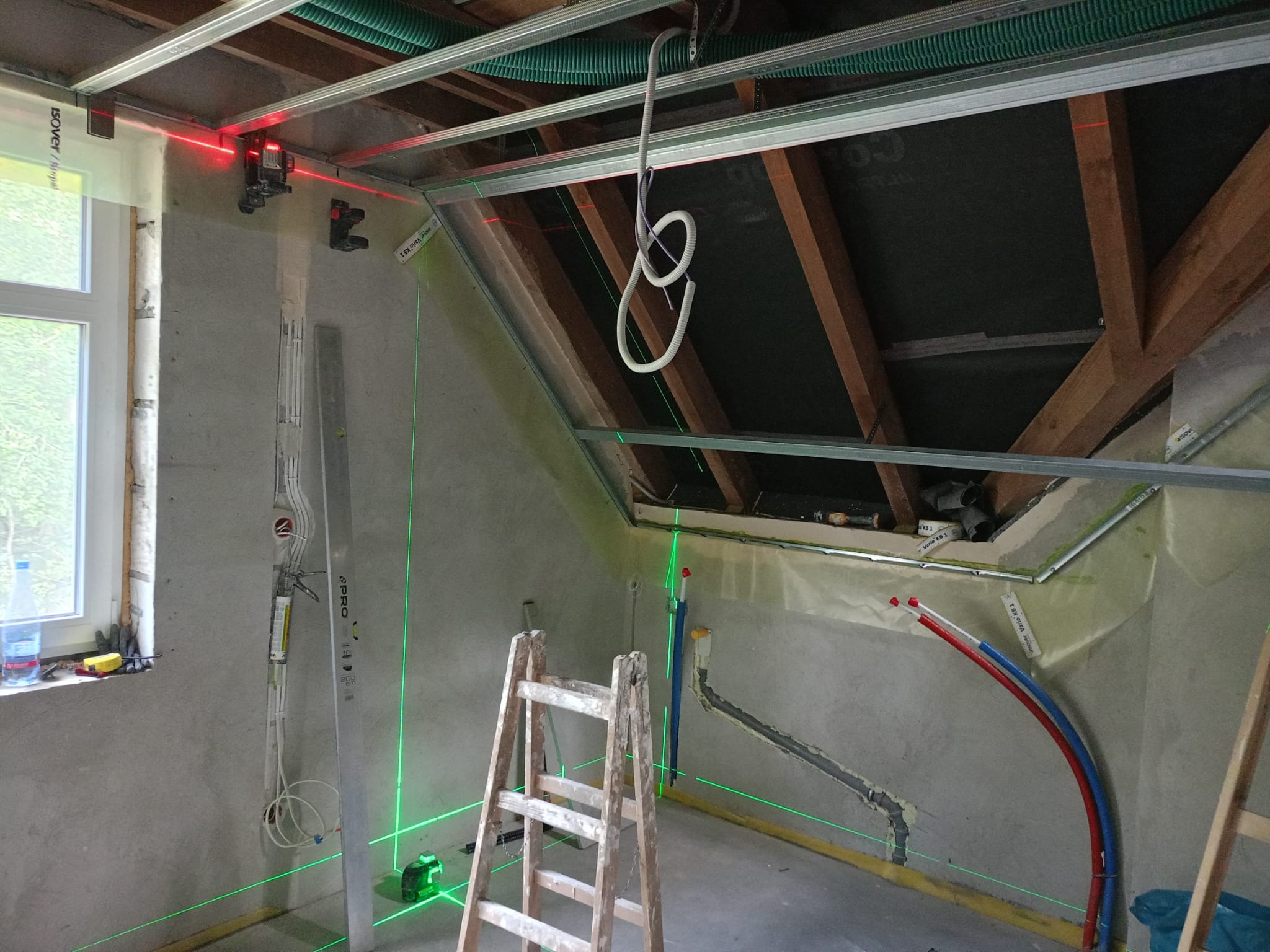 Poddasze w trakcie adaptacji: widoczne profile sufitowe, instalacje, poziomica laserowa wyznaczająca płaszczyzny. Prace wykończeniowe w toku, surowy stan ścian.