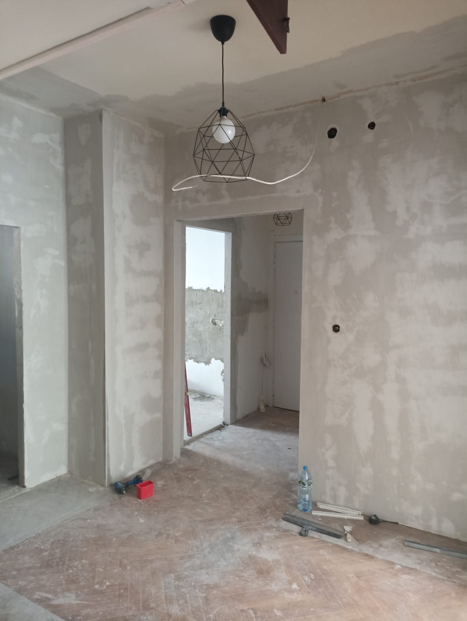 Wnętrze w trakcie szpachlowania: surowe ściany, drewniana podłoga w jodełkę, wisząca lampa z geometrycznym kloszem, narzędzia i butelka wody na podłodze.