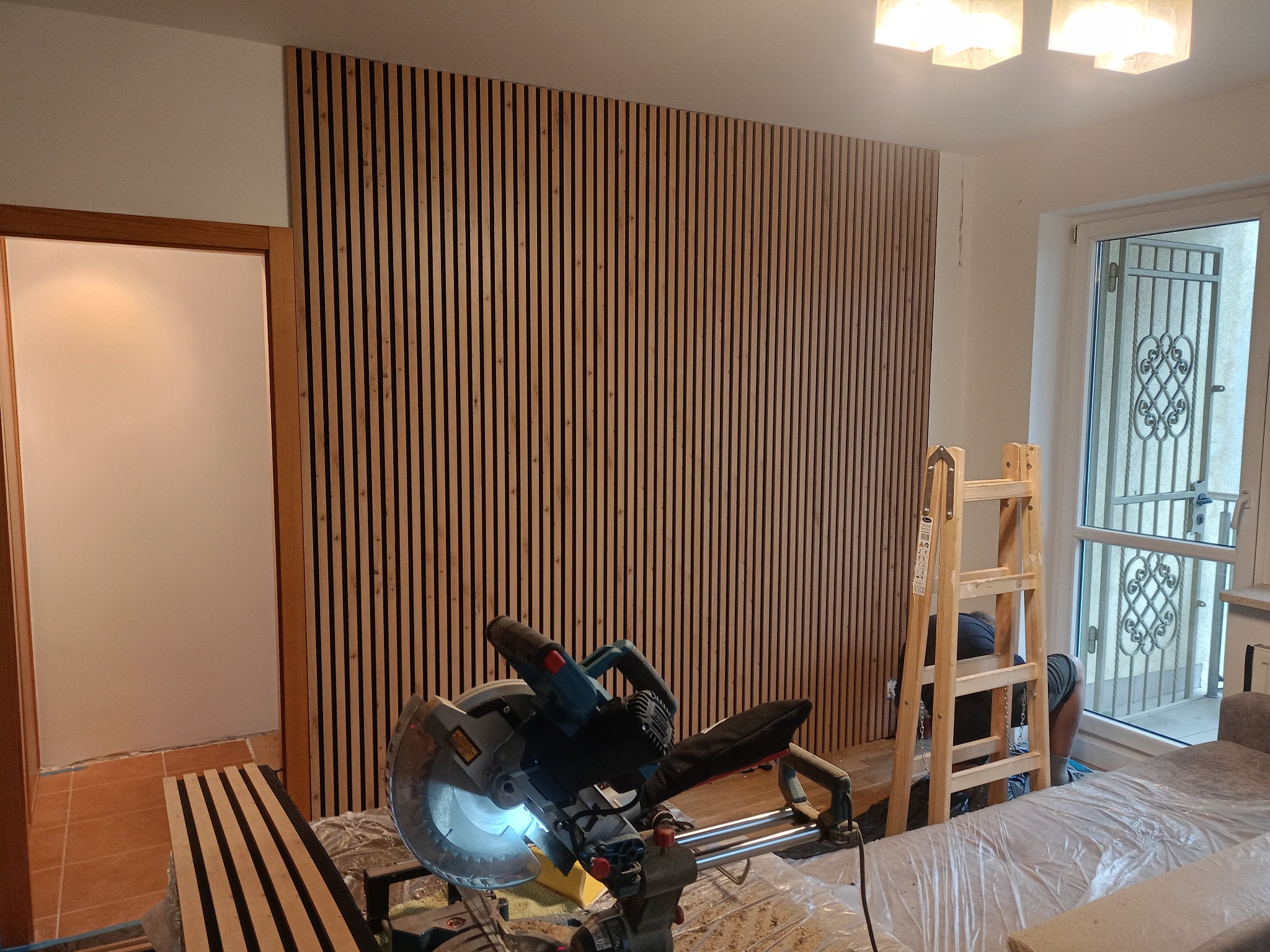 Montaż drewnianych lameli na ścianie, widoczna piła ukośnica, drabina i częściowo zasłonięta osoba. W tle drzwi balkonowe i wejście do innego pomieszczenia.