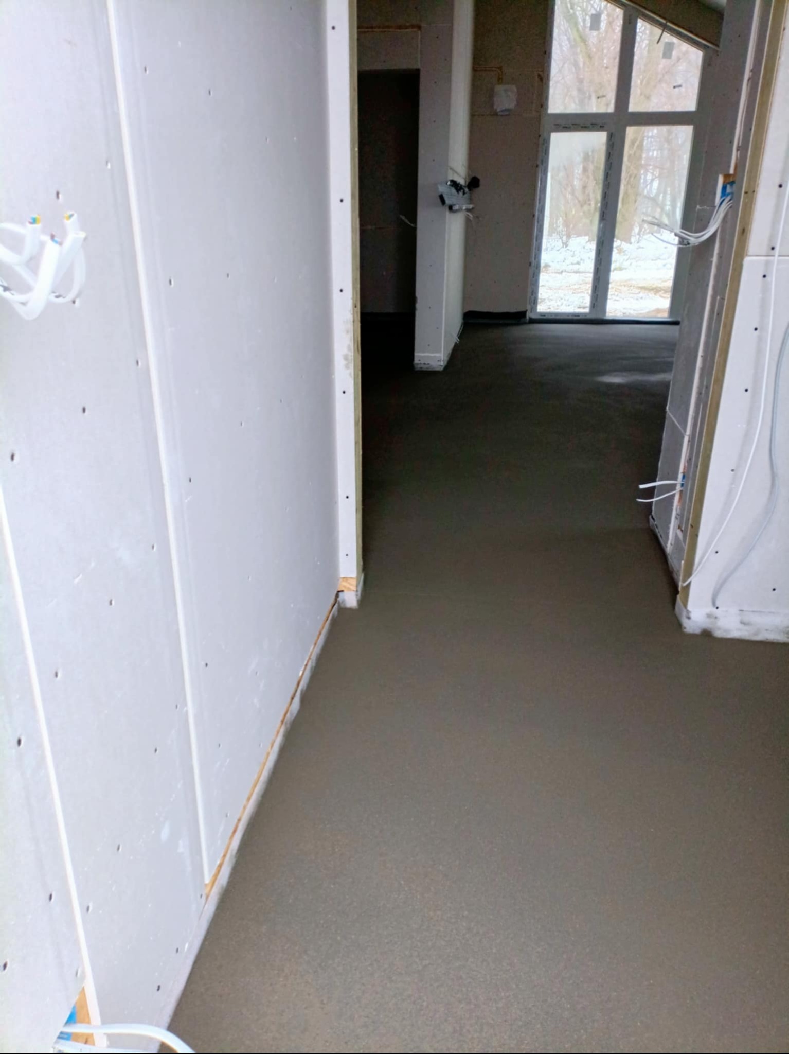 Świeżo wylana, gładka posadzka w jasnym wnętrzu z oknem. Widoczne kable elektryczne przy ścianach, surowy stan wykończenia. Minimalistyczny design, naturalne światło.