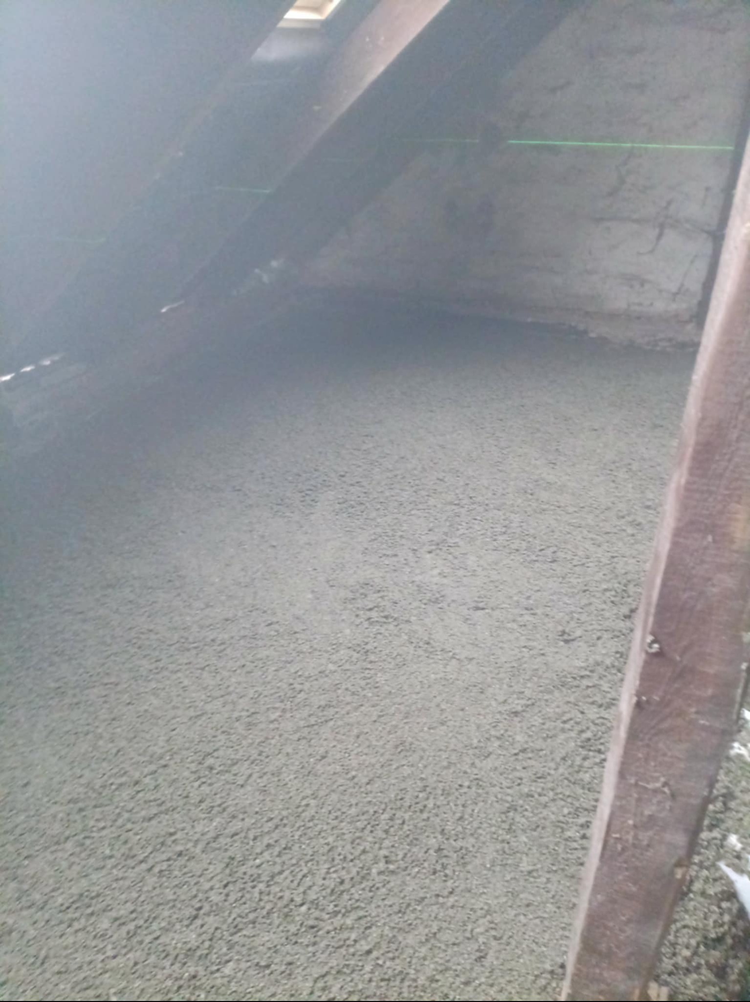 Świeżo wylana, szara posadzka w surowym wnętrzu poddasza z widoczną drewnianą konstrukcją dachu i zieloną linią lasera na ścianie. Widok z góry.