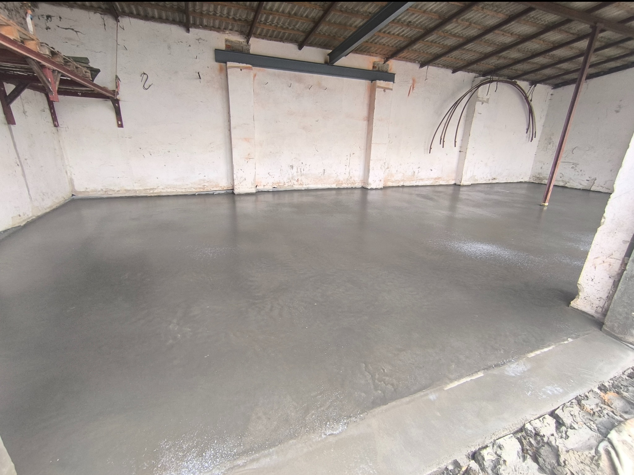 Świeżo wylana, gładka posadzka jastrychowa w surowym wnętrzu z widoczną konstrukcją dachu i ścianami z ubytkami tynku. Widoczne metalowe elementy konstrukcyjne.