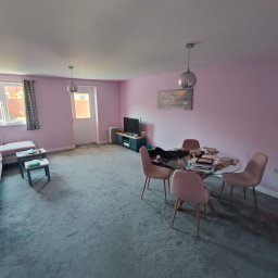 DM Renovations - Jasny salon z różowymi ścianami, szarym dywanem i okrągłym stołem z krzesłami. Widoczne okno z zasłonami, sofa i szafka RTV. Całość w stylu nowoczesnym.