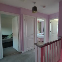 DM Renovations - Korytarz po malowaniu na liliowo z trzema białymi drzwiami i lampą. Widok na pokoje z łóżkami. Balustrada schodów w paski.