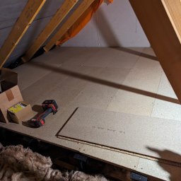 DM Renovations - Adaptacja poddasza: płyty OSB ułożone na podłodze strychu, widoczne krokwie, wkrętarka i karton z wkrętami. Ocieplenie z wełny mineralnej.