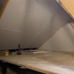 DM Renovations - Płyty gipsowe zamontowane na poddaszu, widoczne drewniane belki konstrukcyjne, wiertarka i karton z wkrętami na podłodze. Prace wykończeniowe na poddaszu.