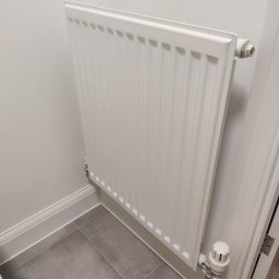 DM Renovations - Biały, nowy grzejnik panelowy zamontowany na białej ścianie w łazience z szarymi płytkami podłogowymi. Widoczny zawór termostatyczny i listwa przypodłogowa.