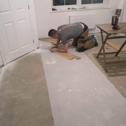 DM Renovations - Parkieciarz na kolanach układa drewniane panele podłogowe na podkładzie w jasnym pokoju z oknem. Na stoliku narzędzia.