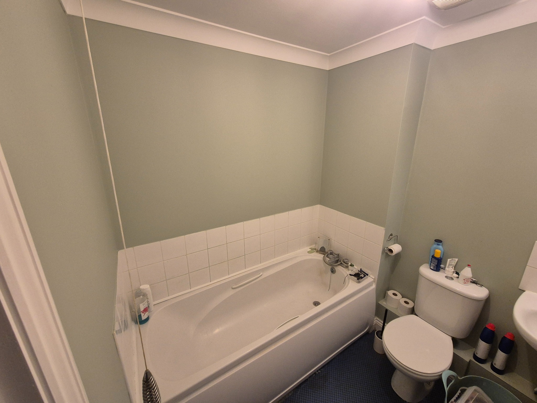 Jasna łazienka z wanną obłożoną białymi płytkami, toaletą i umywalką. Ściany pomalowane na stonowany, zielonkawy kolor. Widoczne akcesoria łazienkowe.