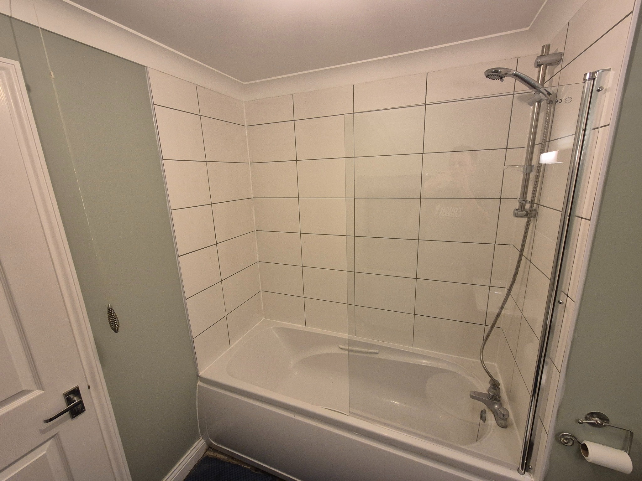 Biała wanna z kabiną prysznicową i białymi płytkami w kwadraty. Widoczny chromowany zestaw prysznicowy i drzwi kabiny. Ściany w odcieniach szarości.