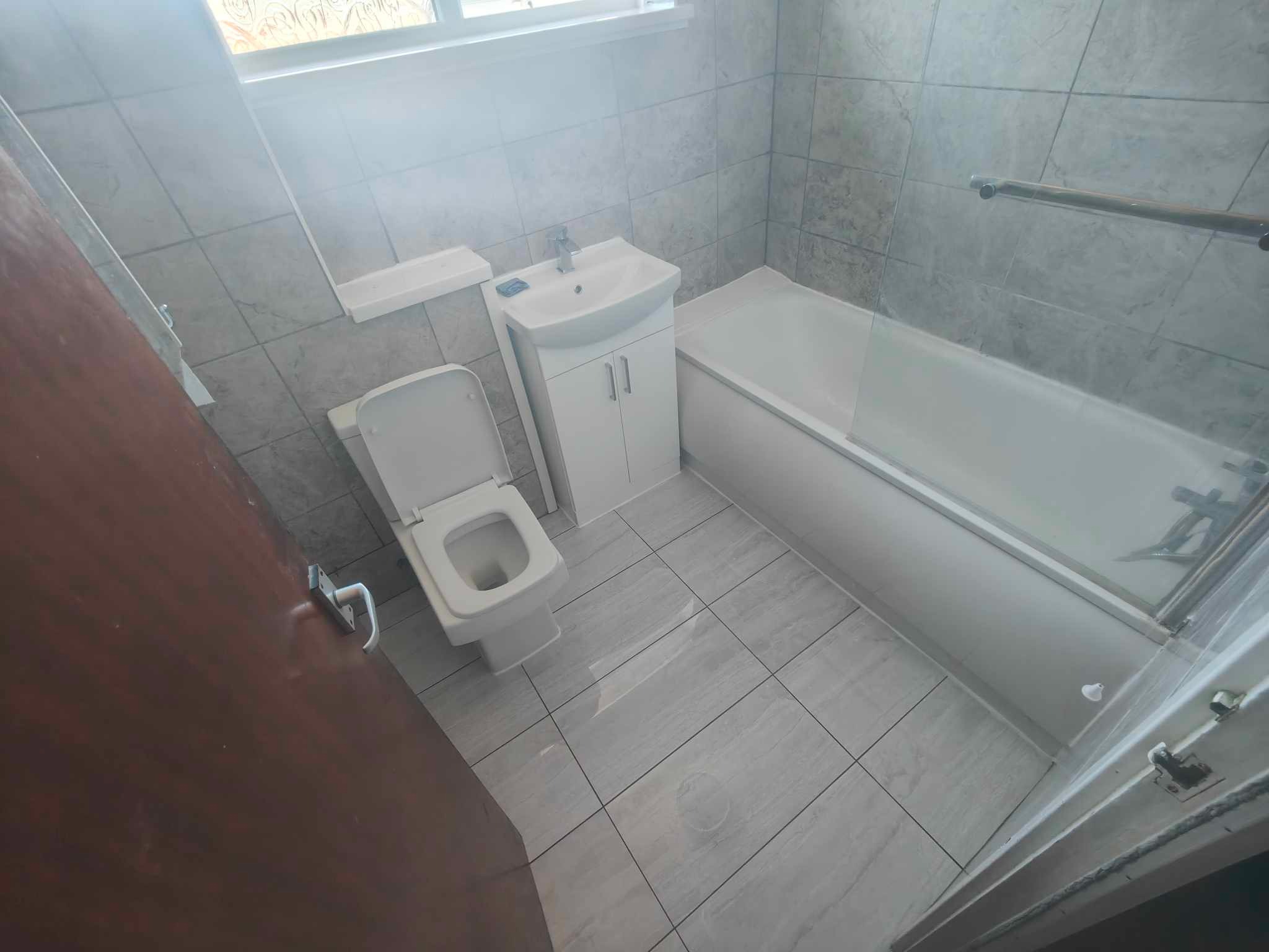 Jasna łazienka po remoncie: biała wanna z parawanem, umywalka z szafką, toaleta i szare płytki imitujące marmur na ścianach i podłodze. Widok zza uchylonych drzwi.