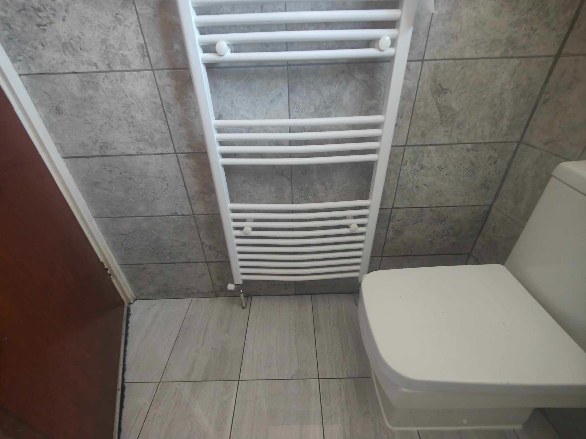 Biały grzejnik drabinkowy w łazience z szarymi płytkami imitującymi marmur na ścianach i podłodze, widoczny fragment toalety. Ujęcie z góry.