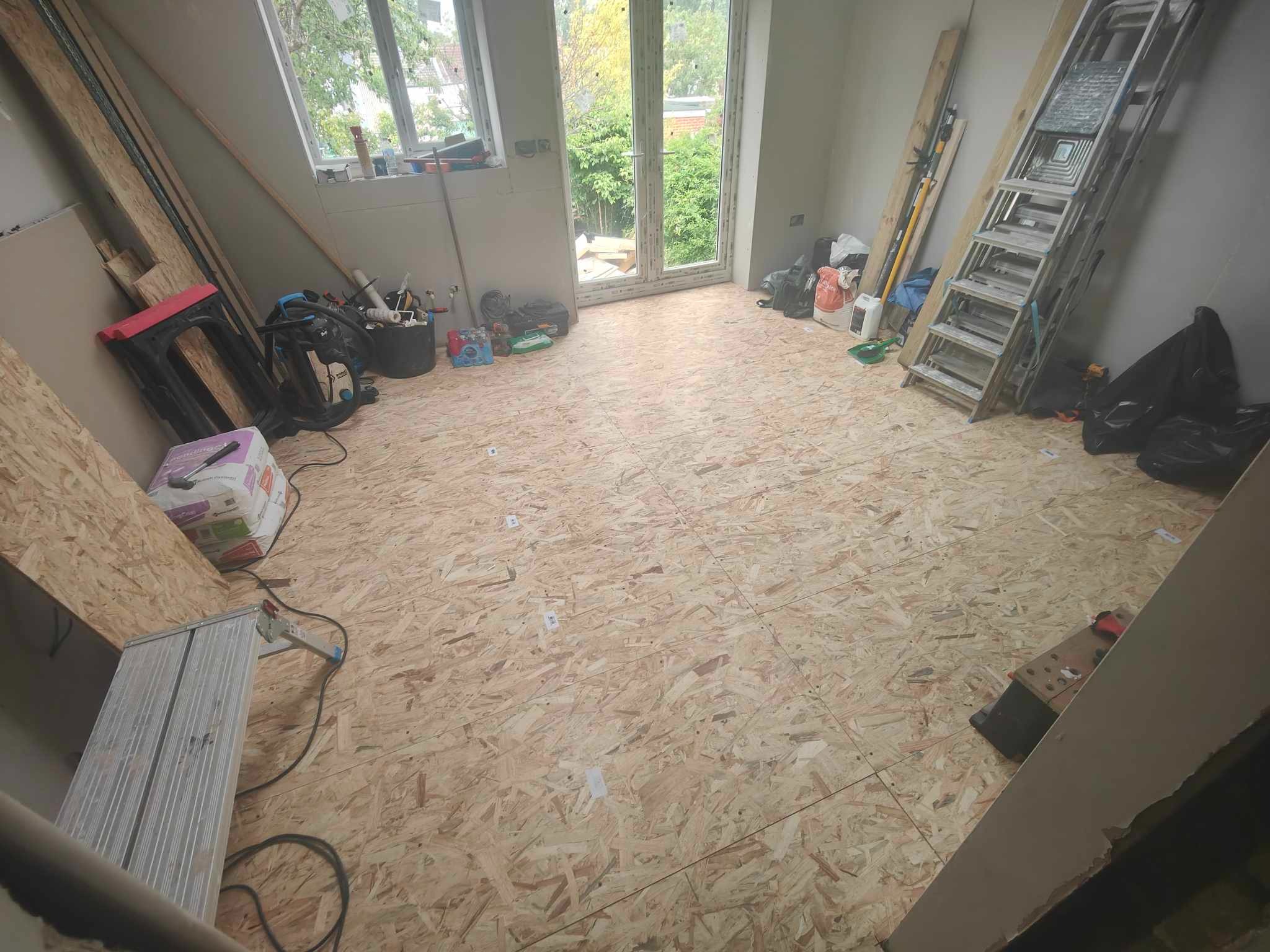 Wnętrze pokoju z ułożoną podłogą z płyt OSB, widoczne narzędzia i drabina. Przygotowanie do dalszych prac wykończeniowych, okno balkonowe z widokiem na zieleń.