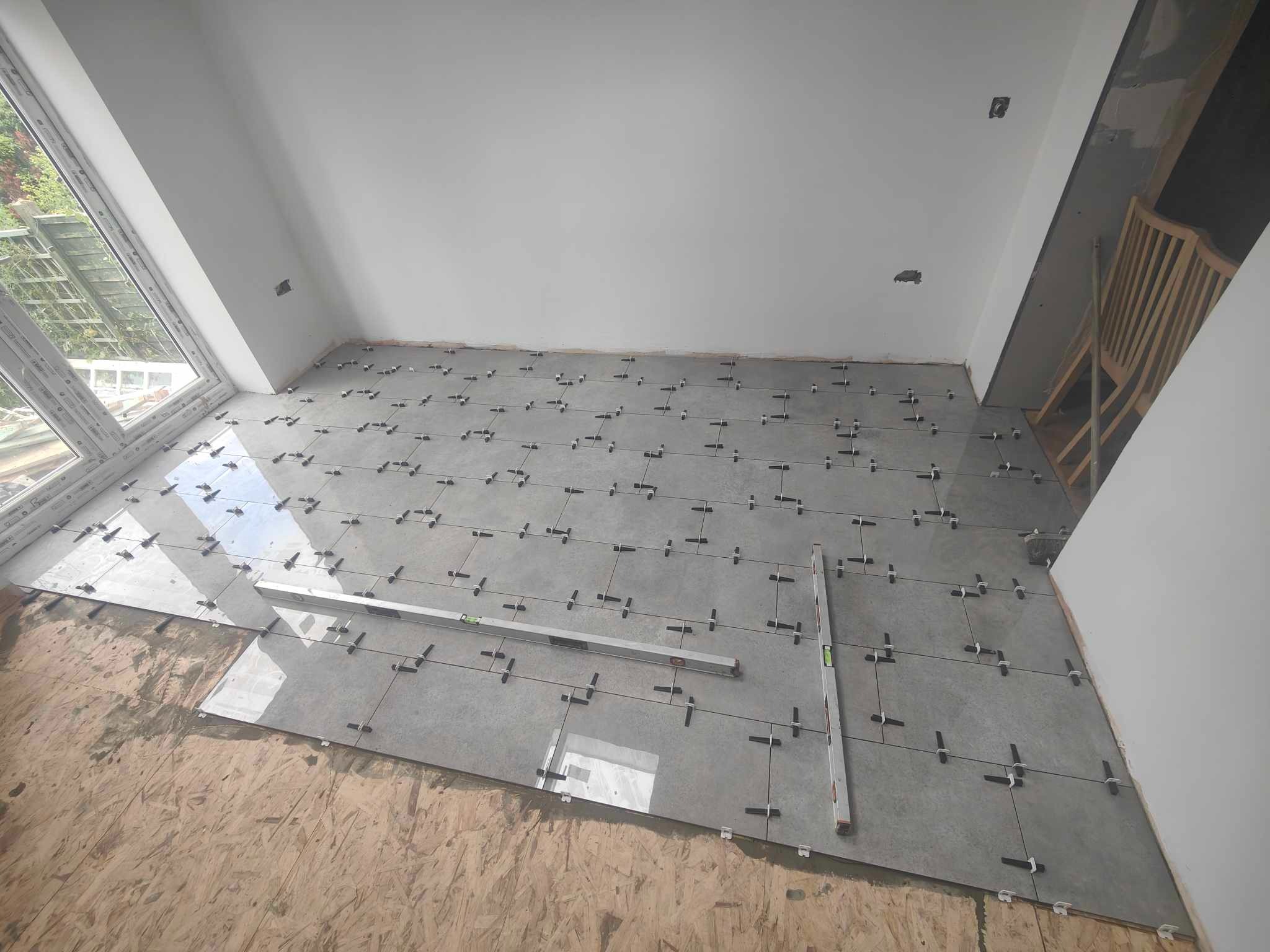 Układanie szarych płytek podłogowych z użyciem poziomicy i krzyżyków dystansowych w jasnym pomieszczeniu z oknem. Widoczna surowa podłoga i fragment schodów.