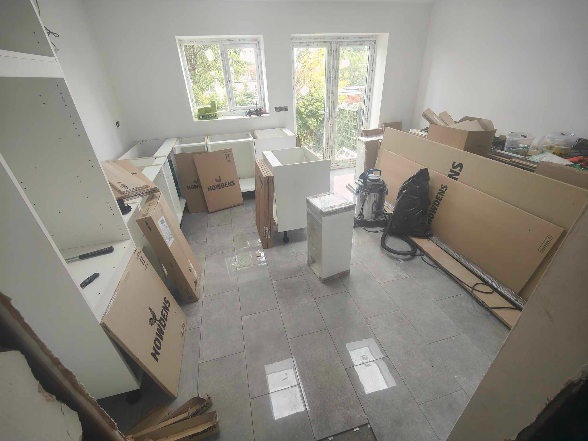 Wnętrze w trakcie montażu kuchni: białe szafki, kartony Howdens, szare płytki podłogowe, okna z widokiem na zieleń. Etap prac przed finalnym wykończeniem.