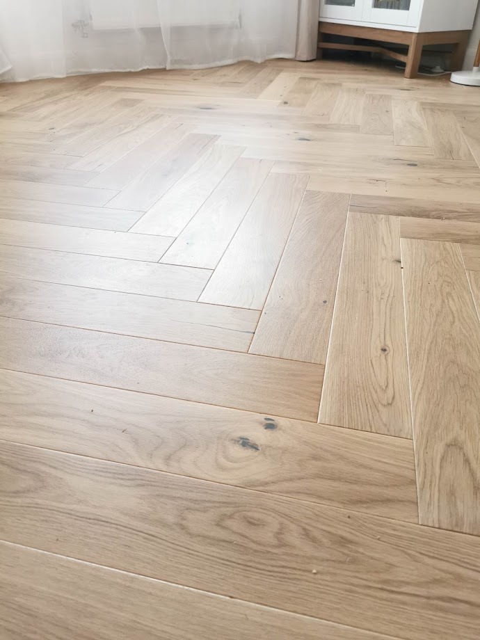 Jasna, dębowa podłoga ułożona w jodełkę francuską, z widocznymi słojami drewna i subtelnymi przebarwieniami, w tle fragment białej szafki i zasłony.