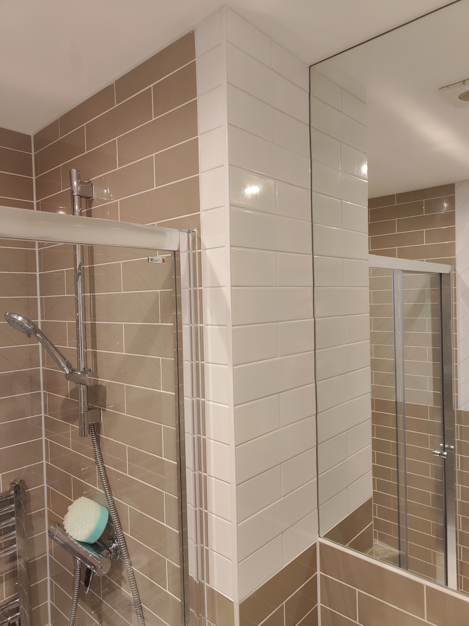 Wykończona kabina prysznicowa z beżowymi i białymi, prostokątnymi płytkami. Widoczne detale: chromowana armatura, szklane drzwi kabiny i odbicie w lustrze.