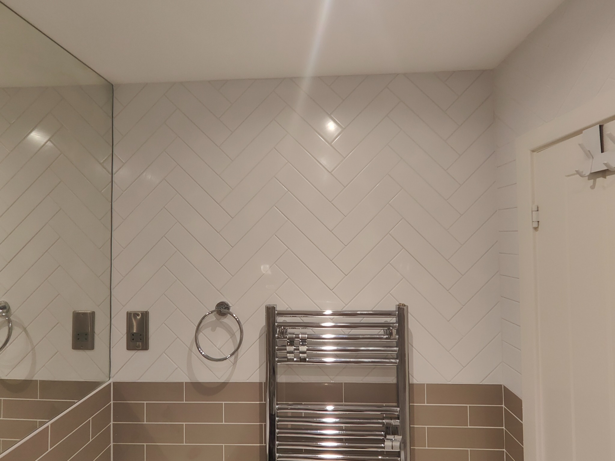 Białe, błyszczące płytki ułożone w jodełkę na ścianie łazienki, z chromowanym grzejnikiem drabinkowym i okrągłym wieszakiem na ręcznik. Widoczne gniazdko elektryczne i lustro.