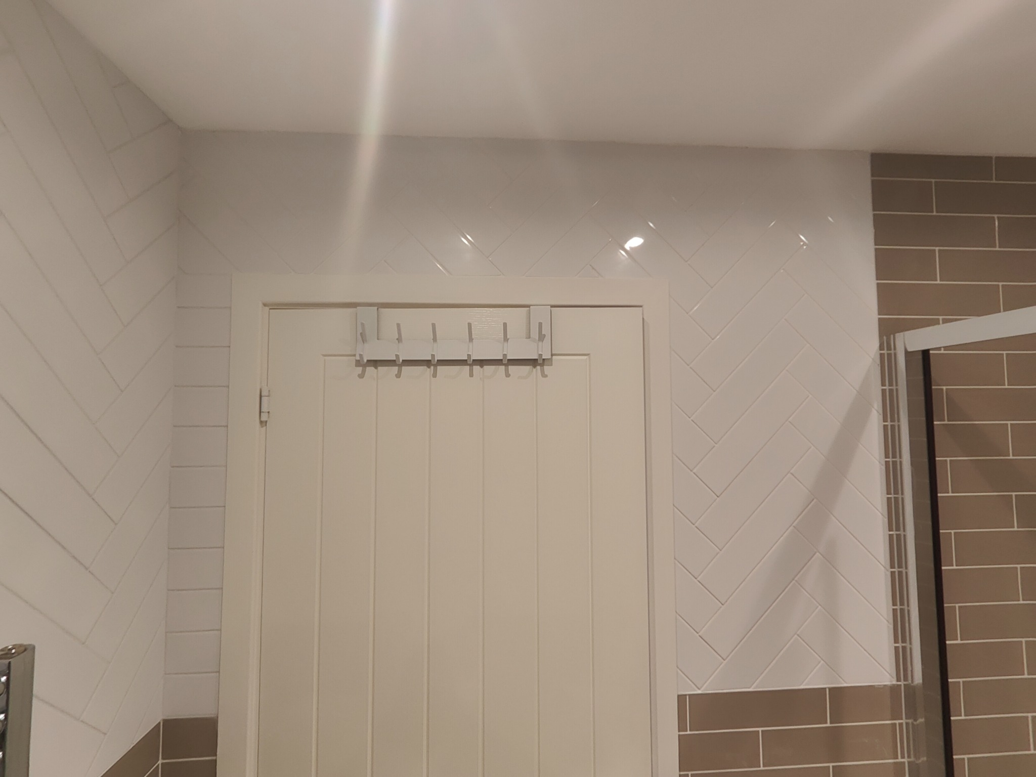 Białe płytki ułożone w jodełkę na ścianie łazienki, obok drzwi z wieszakiem. Dolny pas ściany wykończony brązowymi płytkami.