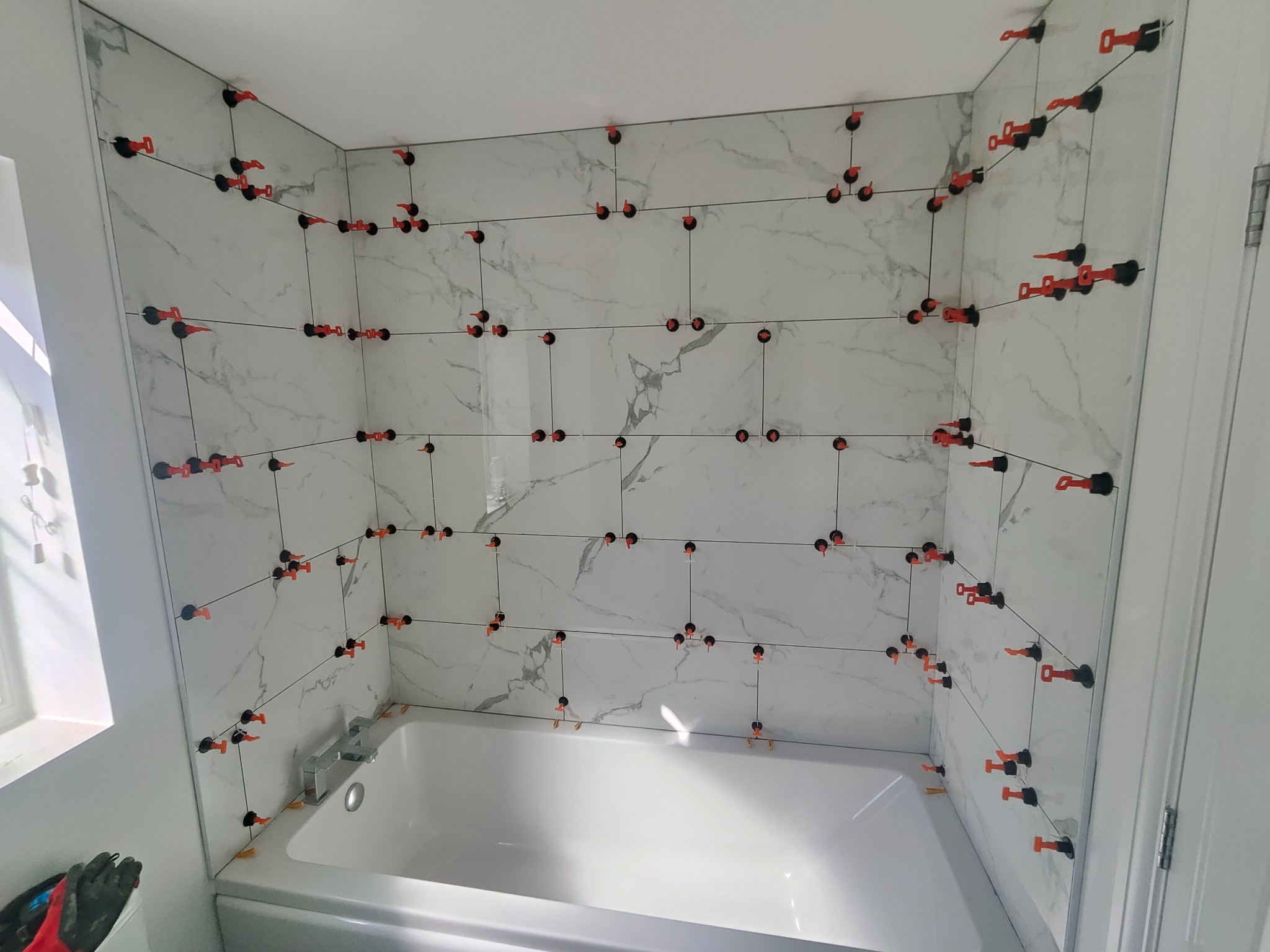 Białe płytki imitujące marmur na ścianie nad wanną, z systemem poziomowania płytek (klipsy i kliny) w trakcie montażu. Widok na fragment łazienki.