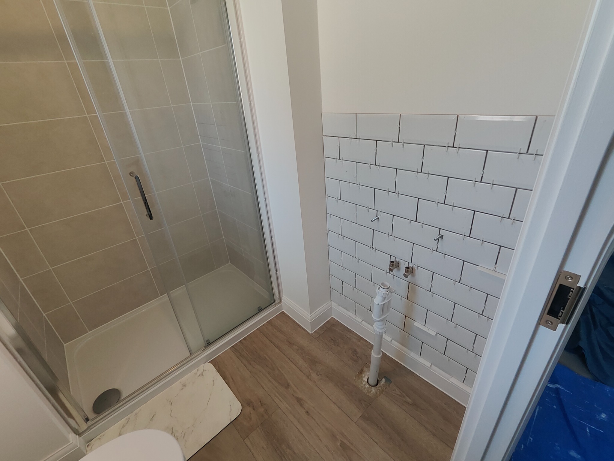 Łazienka w trakcie remontu: kabina prysznicowa, drewnopodobna podłoga i białe płytki na ścianie, częściowo ułożone z krzyżykami dystansowymi. Widoczne rury i otwarta przestrzeń pod umywalkę.