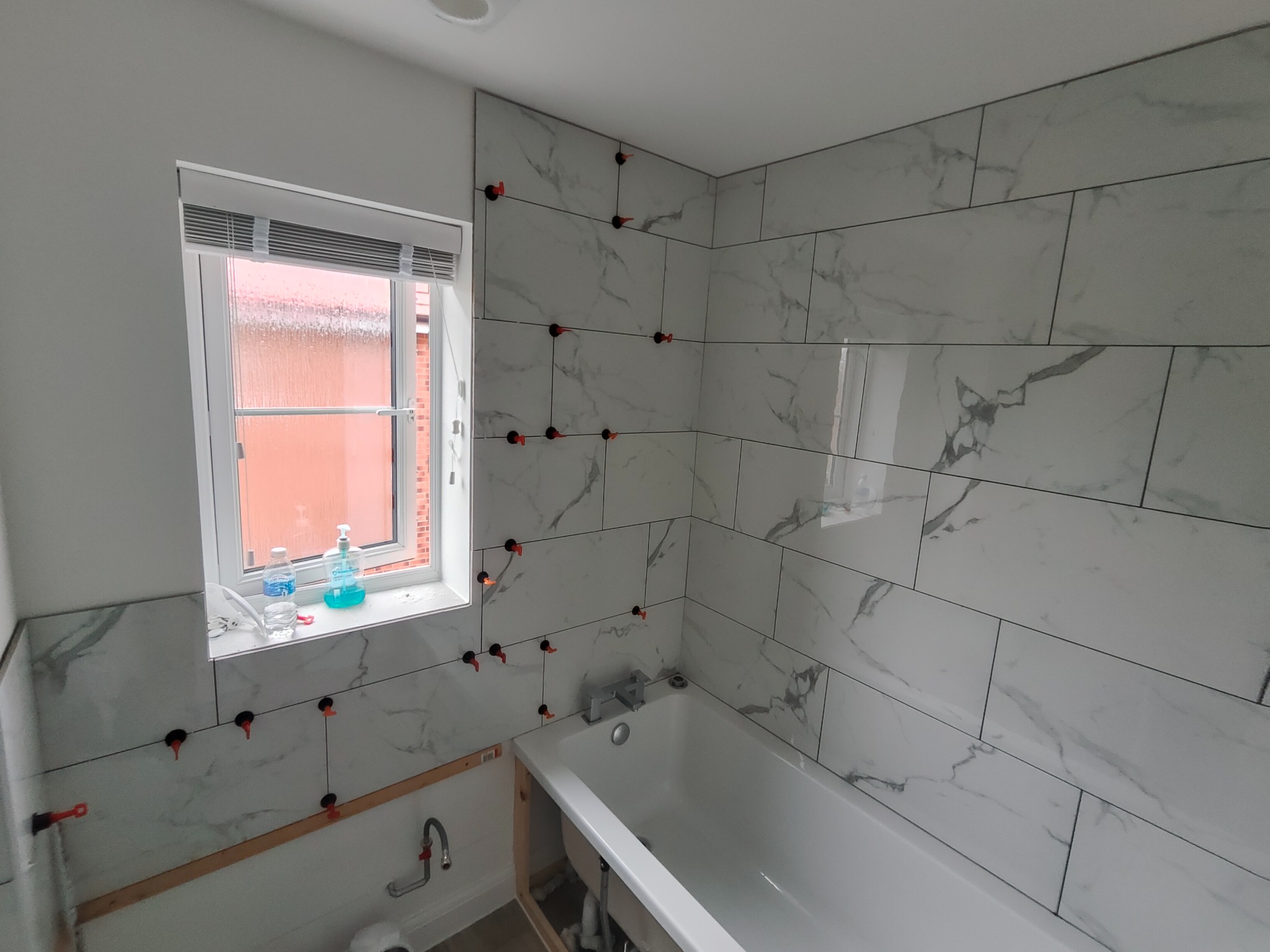 Białe płytki imitujące marmur, ułożone w łazience, z widocznymi krzyżykami dystansowymi. Okno z roletami i butelki na parapecie. Wanna w rogu.