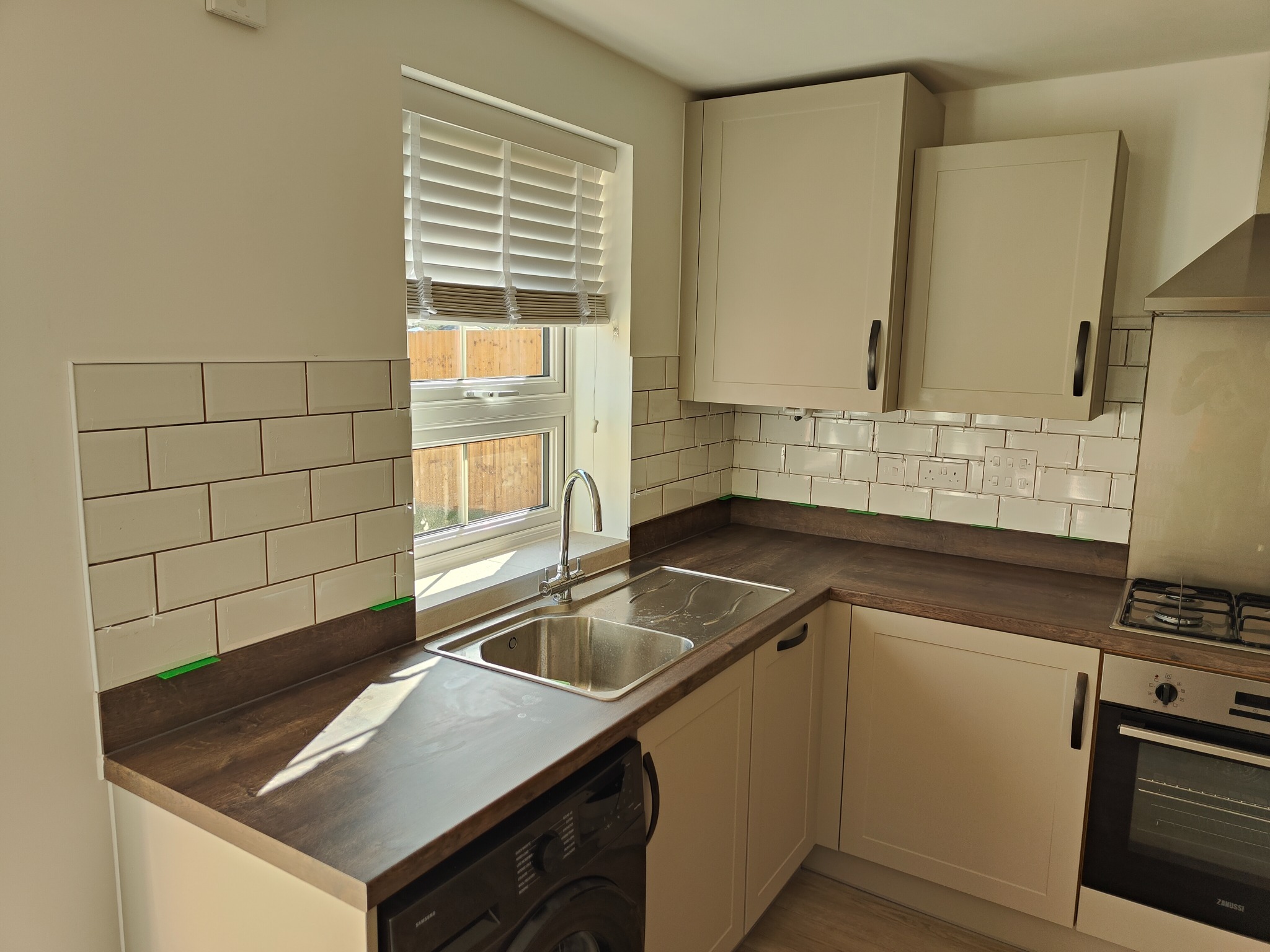 Jasna kuchnia po remoncie: kremowe szafki, ciemny blat, białe płytki na ścianie. Widoczny zlew, okno z żaluzjami i sprzęt AGD. Świeżo położone płytki z zachowanymi odstępami.