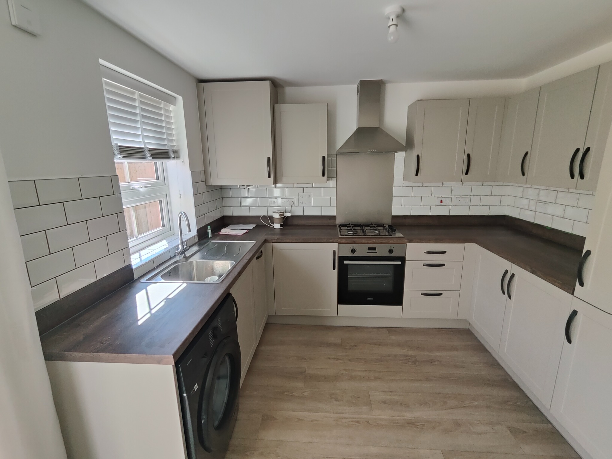 Jasna kuchnia po remoncie: białe szafki z ciemnymi uchwytami, drewniany blat, białe płytki na ścianie, okno z żaluzjami, sprzęt AGD. Nowoczesny design.