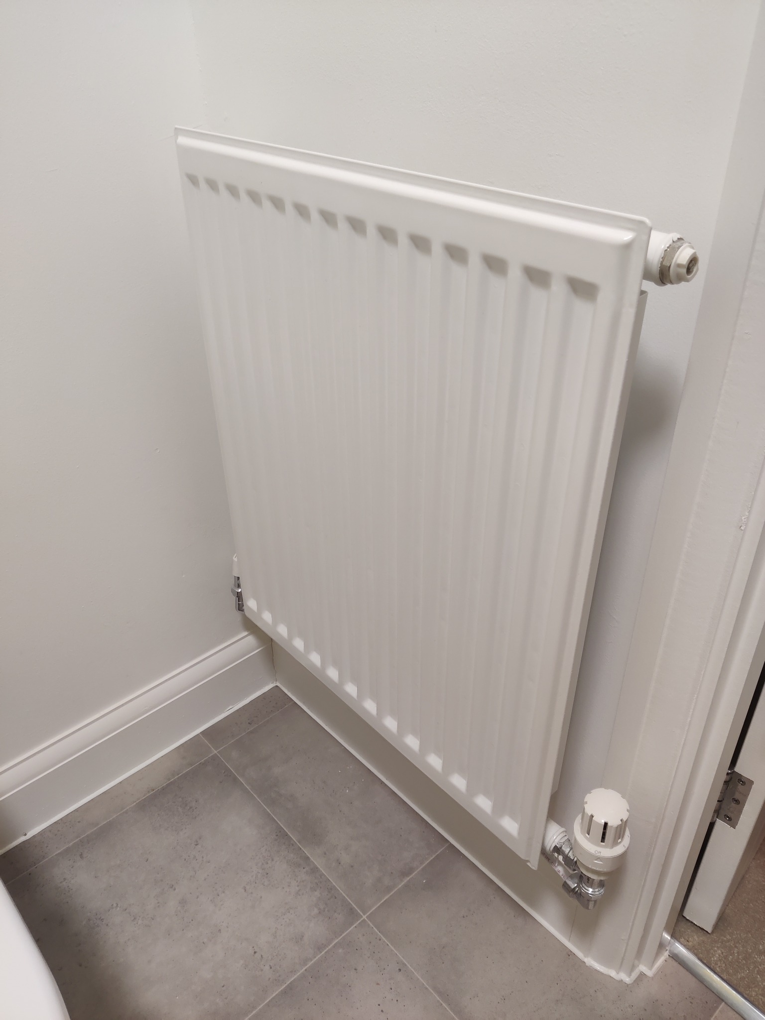 Biały, nowy grzejnik panelowy zamontowany na białej ścianie w łazience z szarymi płytkami podłogowymi. Widoczny zawór termostatyczny i listwa przypodłogowa.
