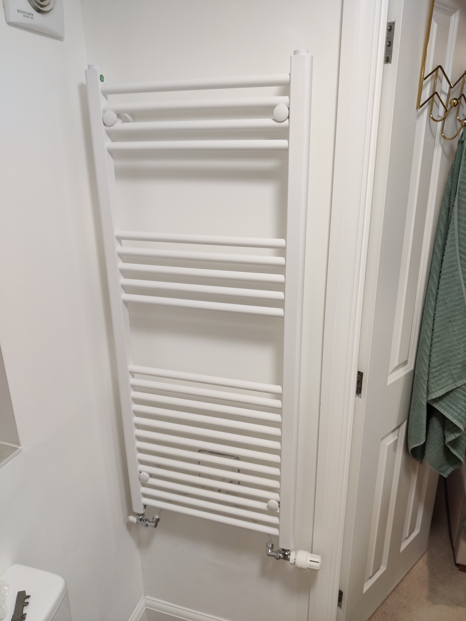 Biały, drabinkowy grzejnik łazienkowy zamontowany na ścianie, z widocznymi zaworami i fragmentem drzwi z wieszakiem na ręcznik. Minimalistyczny design, jasne wnętrze.